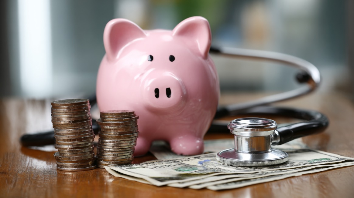 A pink piggy bank with coins, cash, and a stethoscope representing health insurance savings.